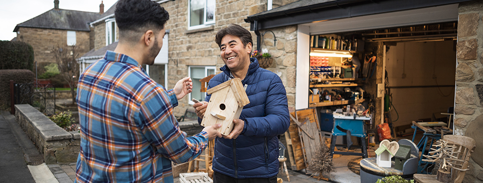 A smiling man, standing outside of a residential garage workshop, displaying a handmade wooden birdhouse to a potential customer.