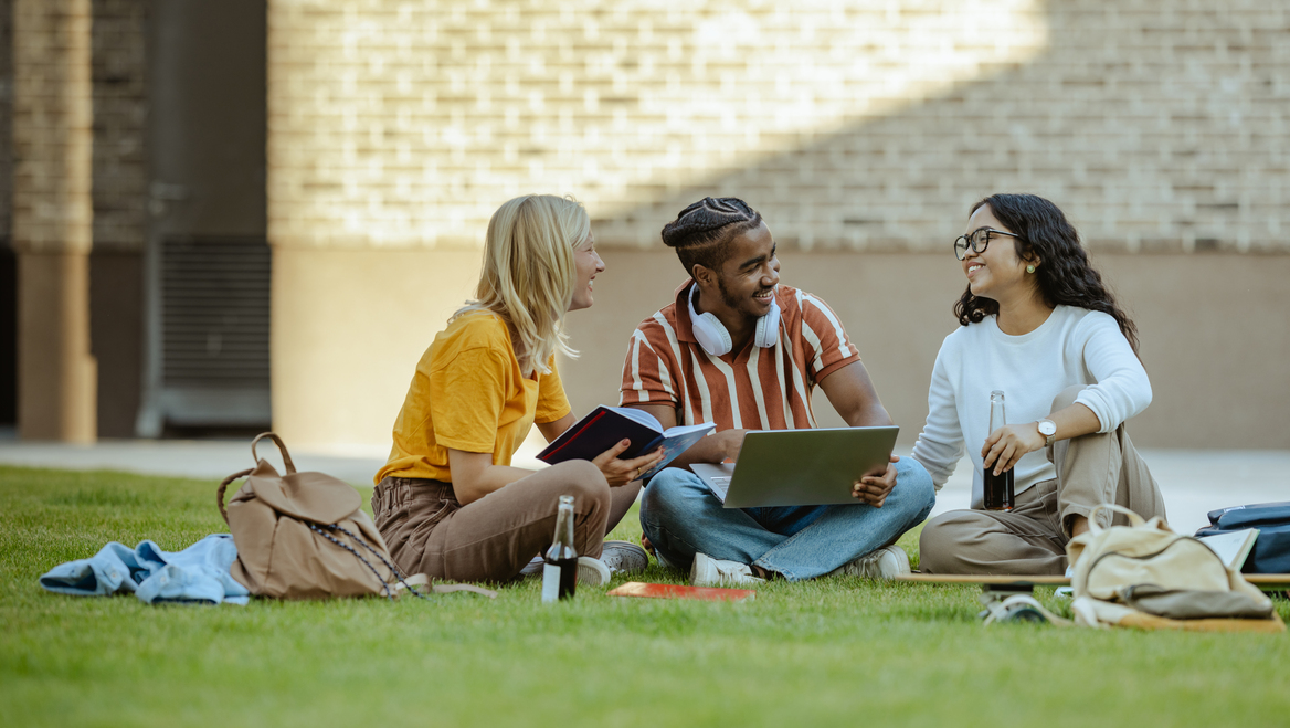 Three students sit on the grass on a college campus.