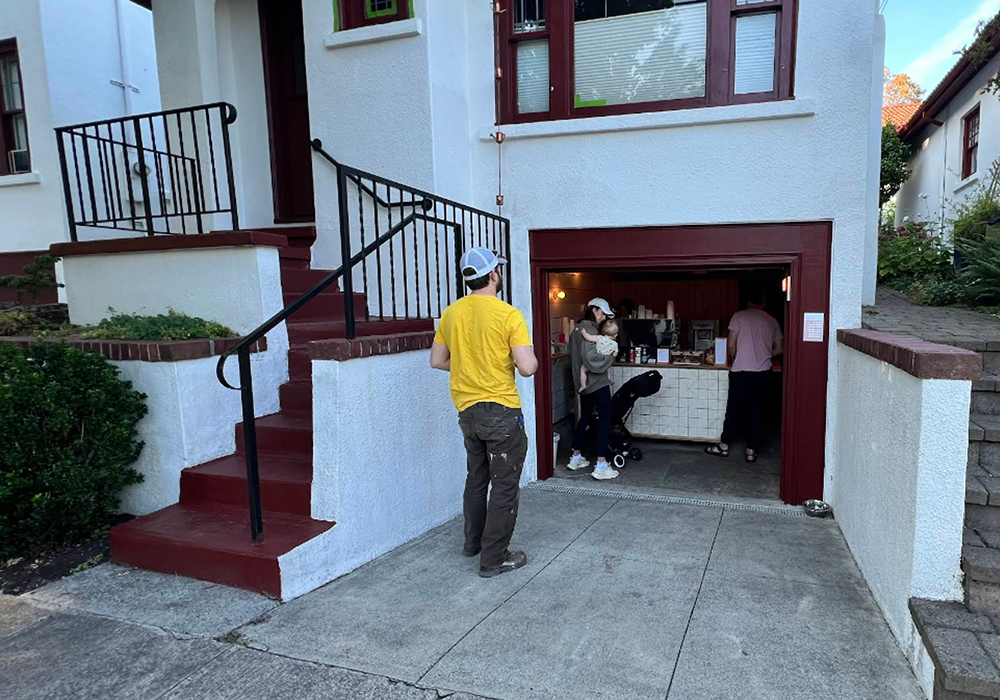 A man waits outside a converted single-car, tuck-under garage, while a young mother holding a baby orders a coffee at the counter inside.