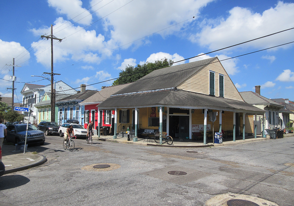 A single-story, siding-clad wooden commercial structure, with a hipped-roof overhang shading the entrance of a store, at the corner of two residential streets lined by modest shotgun homes.