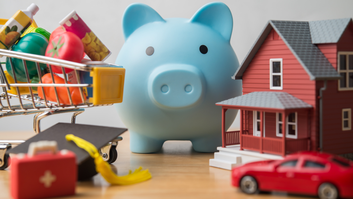 A toy piggy bank, shopping cart, house, and car sit on a table.