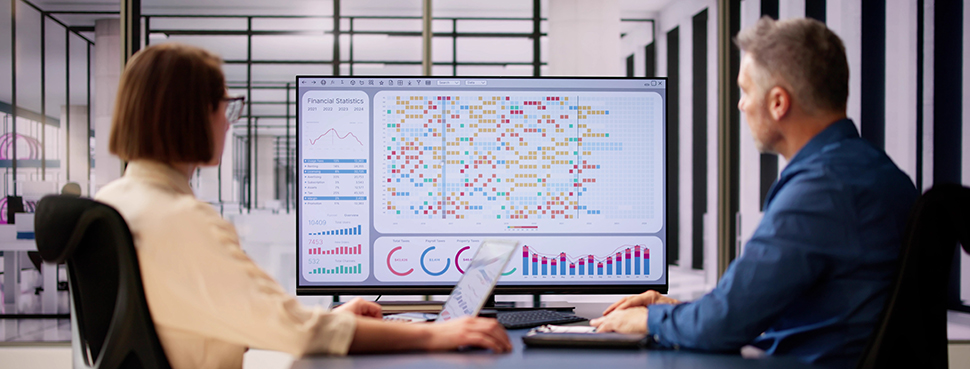 A man and a woman sit in a conference room studying a complex data display on a large central monitor.