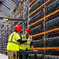 Two technicians in hard hats and high-visibility vests inspect a server rack in a data center.