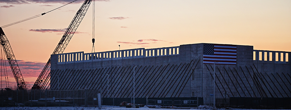 A massive stone-framed structure, draped with a large U.S. flag, with construction cranes in the background.