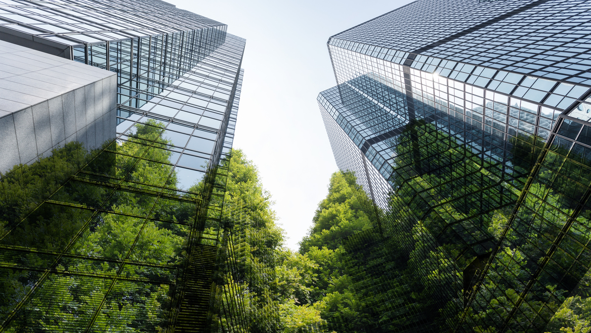 Glass skyscrapers rise into the sky with green trees on the ground.