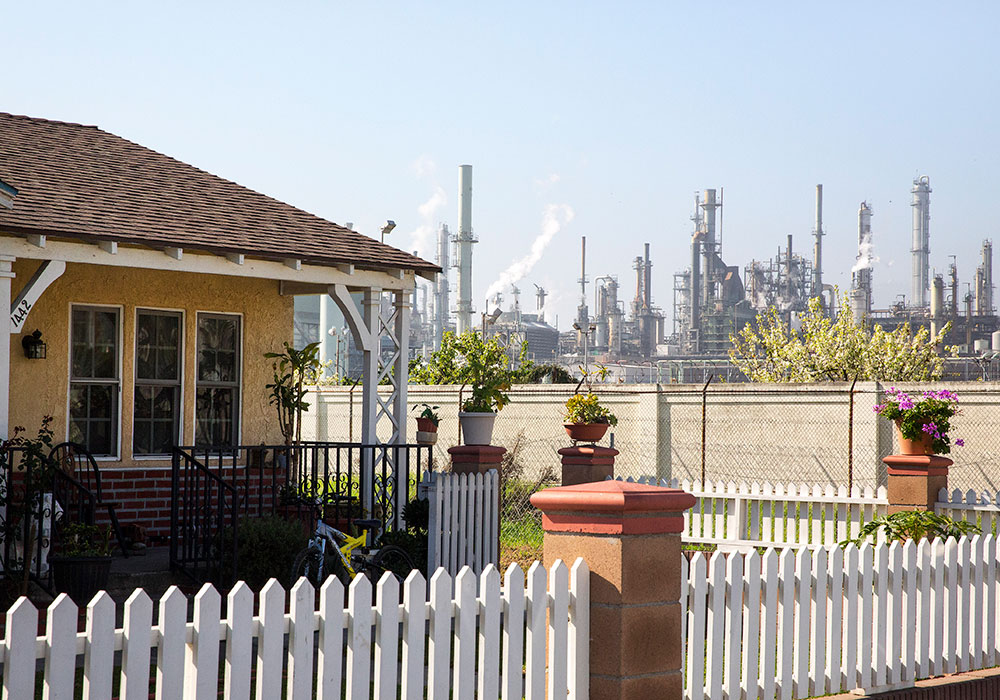 Not far beyond the white picket fence surrounding a home in the Wilmington neighborhood of Los Angeles is one of three area oil refineries — part of a “systemic land-use problem” that exposes residents to hazardous chemicals. Wilmington is working with the Clean Up Green Up program, which focuses on greening polluted neighborhoods. Photo by Monica Almeida/The New York Times.