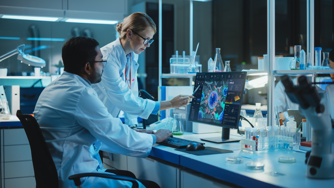 Two people in white coats sit in a lab looking at a computer screen.