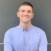 Max Pastore headshot (Zoning Practice December 2025) A confident, smiling person with short hair in glasses and a light colored button-up shirt.