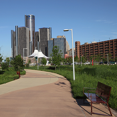Detroit skyline viewed from a paved pathway with a park bench, showcasing modern architecture and green space.