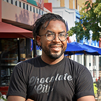 Bobby Boone, AICP, headshot (Zoning Practice December 2025) A person with glasses and short dreadlocks smiles outdoors, wearing a 'Chocolate City' T-shirt, with colorful buildings and trees along a pedestrian-oriented mixed-use street in the background.