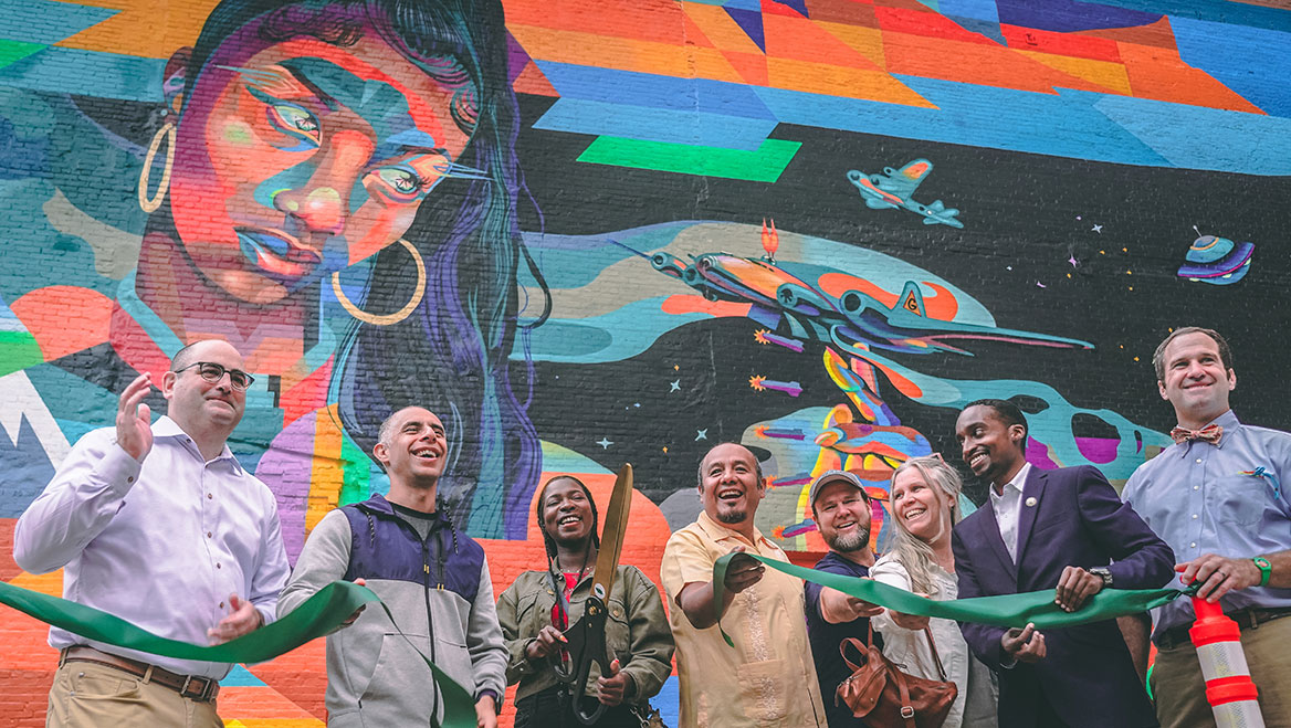Providence Mayor Jorge Elorza (in grey), mural artist Garden of Journey (center, with scissors), and government and tourism officials attend the official dedication of the The Avenue Concept mural “Salt Water" in September 2021. Local members of the community were hired to help the artist complete the work. Photo by David Santilli.