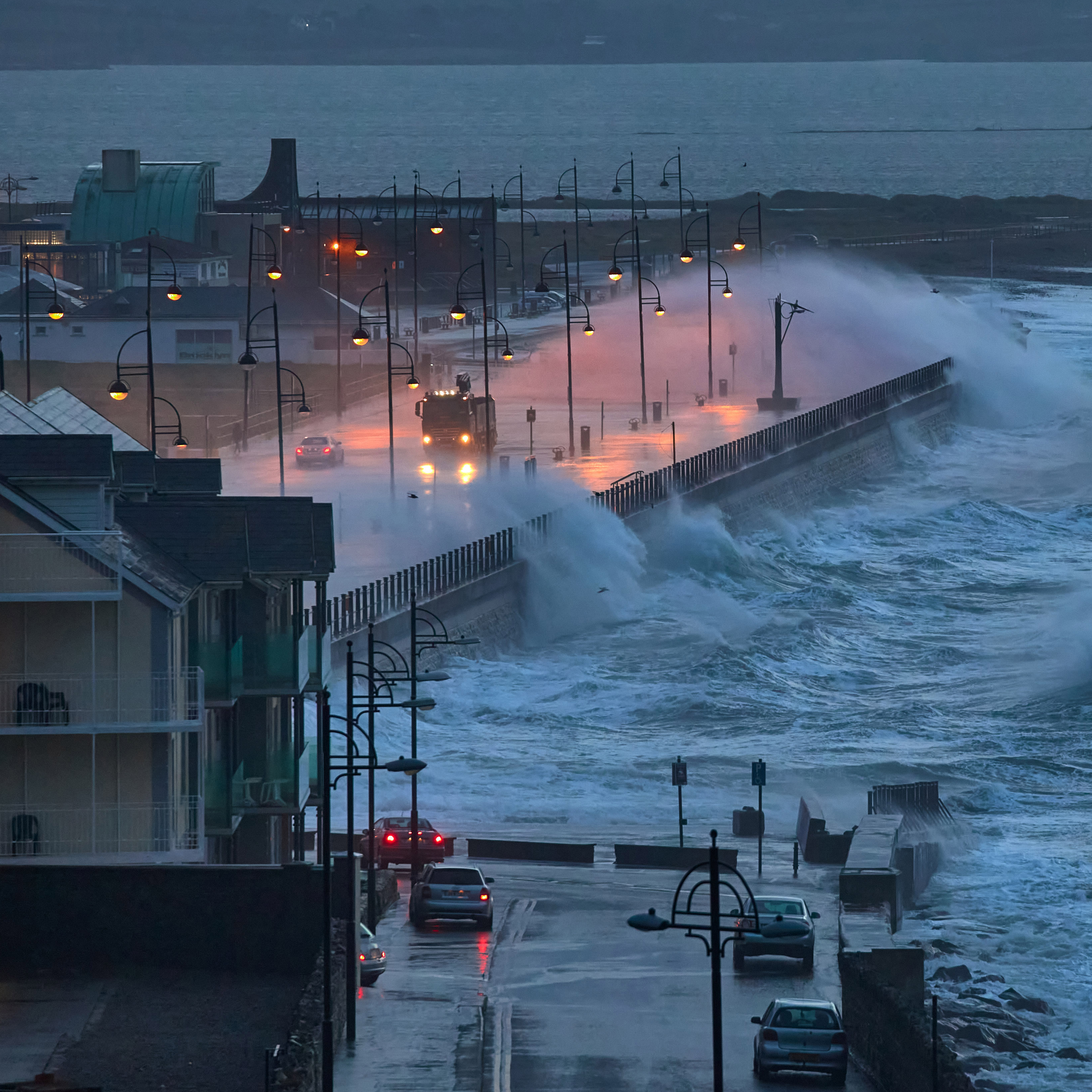 Large waves and high winds batter a coastal district with townhouses and ornamental streetlights, washing over sidewalks and roadways on a stormy evening.