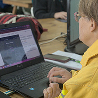 Woman in yellow jacket uses laptop to view wildfire map.