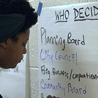 Woman writing "Who Decides?" on a whiteboard, listing Planning Board, City Council, Big Business/corporations, and Community Board.