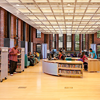 Bright library interior with brick walls, bookshelves, students, and computer stations.