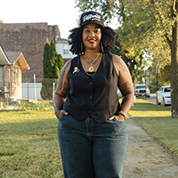 Woman in vest and jeans standing on sidewalk in a residential neighborhood.