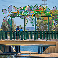 Couple on a bridge admiring a colorful kinetic sculpture in a park.