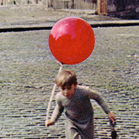 Boy running with a red balloon down a cobblestone street.