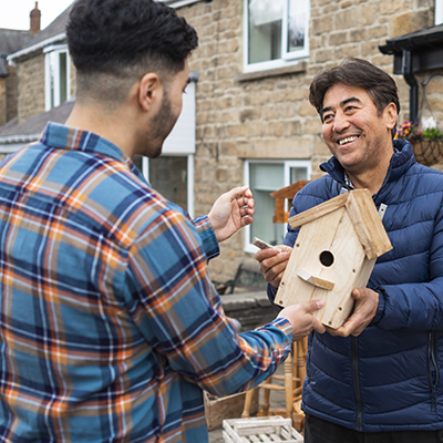 A smiling man, standing outside of a residential garage workshop, displaying a handmade wooden birdhouse to a potential customer.