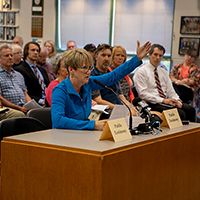 In a meeting room full of people sitting in chairs, a woman with short brown hair and glasses in a blue jacket sits at a table with signs re
