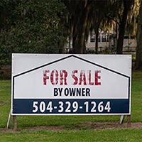 A sign that says For Sale By Owner in a grassy field, with trees in the background