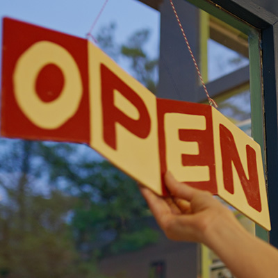 A woman reaches to turn an "OPEN" sign on the door of a storefront business.