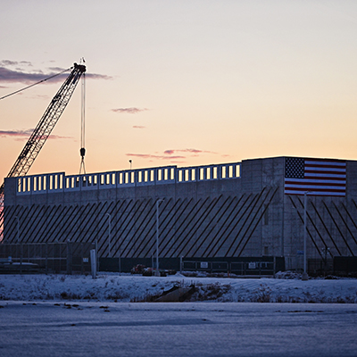 A massive stone-framed structure, draped with a large U.S. flag, with construction cranes in the background.