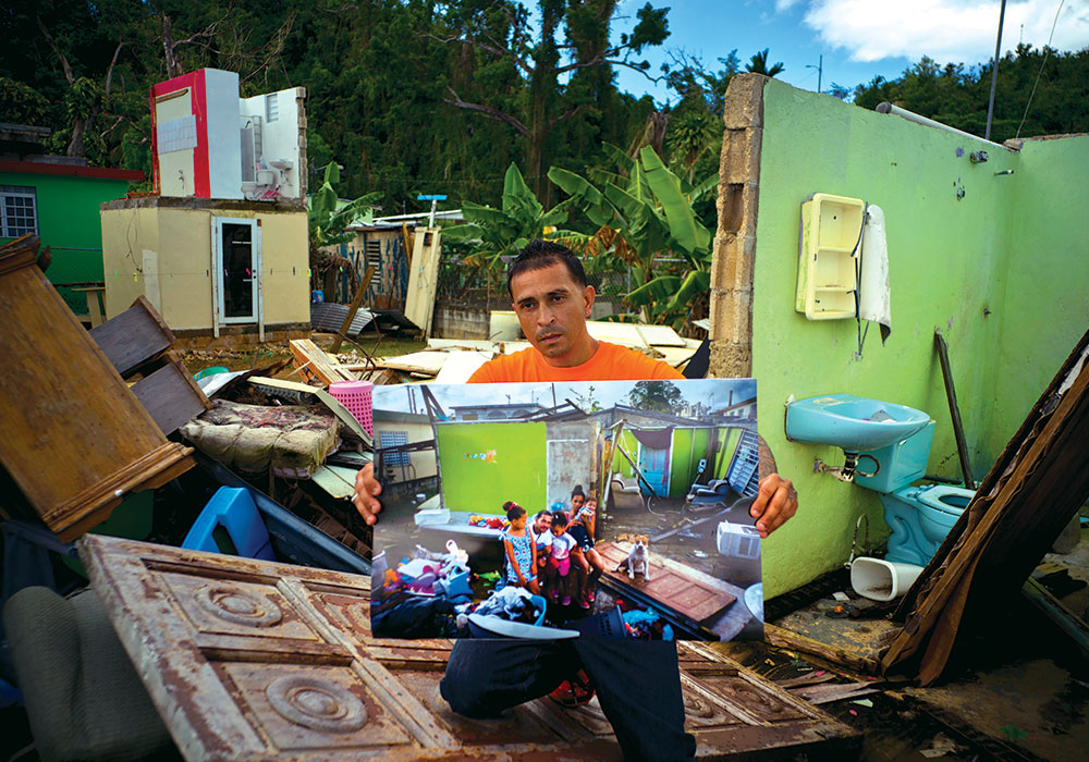 Seven months after his home in Toa Baja was destroyed by Hurricane Maria in October 2017, Arden Dragoni holds a photo of his family taken just after the storm in the same spot. At the time of the 2018 photo, the unemployed construction worker and security guard was separated from his family while his wife and children lived in a FEMA-subsidized apartment and he lived with his father.