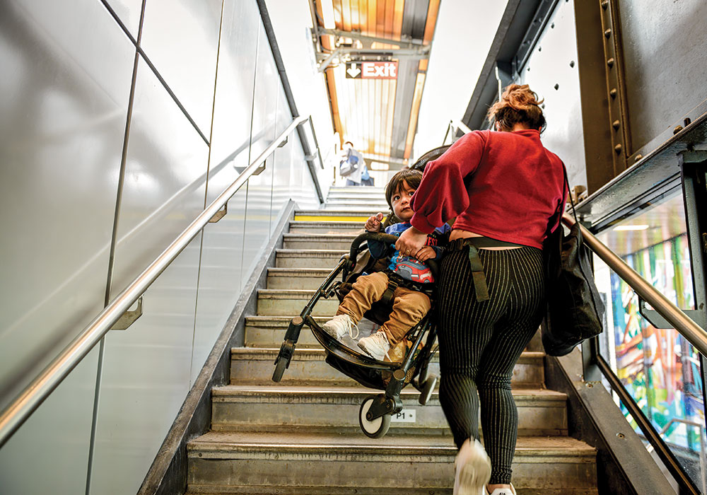 A woman lugs her toddler and stroller up the subway stairs in Queens, New York. Photo by Marian Carrasquero/The New York Times.