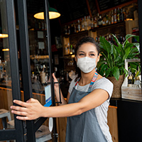 A mask-wearing employee of a small business that's open during the pandemic holds open the door to her shop. Getty Images.